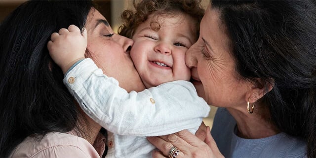 Un niño sonriente abrazado y besado por dos mujeres, una a cada lado.