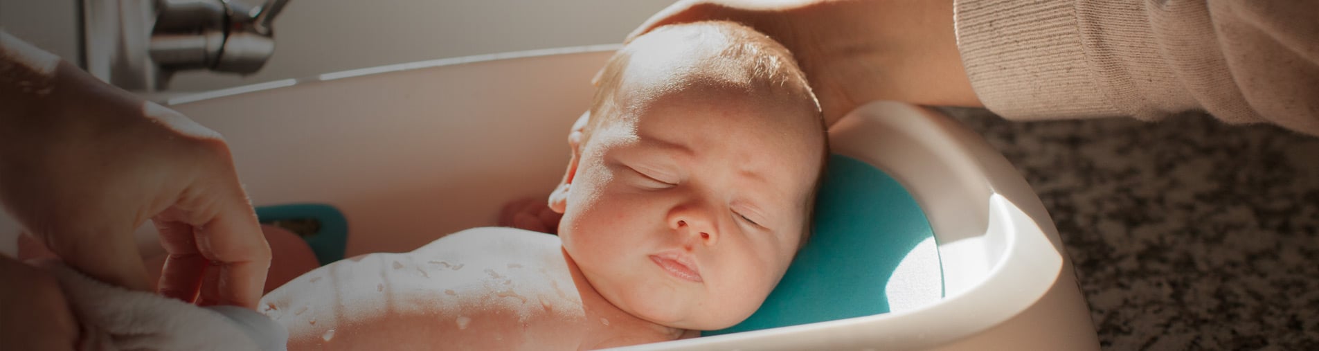 Bebé en una pequeña bañera, siendo lavado suavemente y sostenido por manos adultas, situado en un baño iluminado por el sol con una encimera de granito.