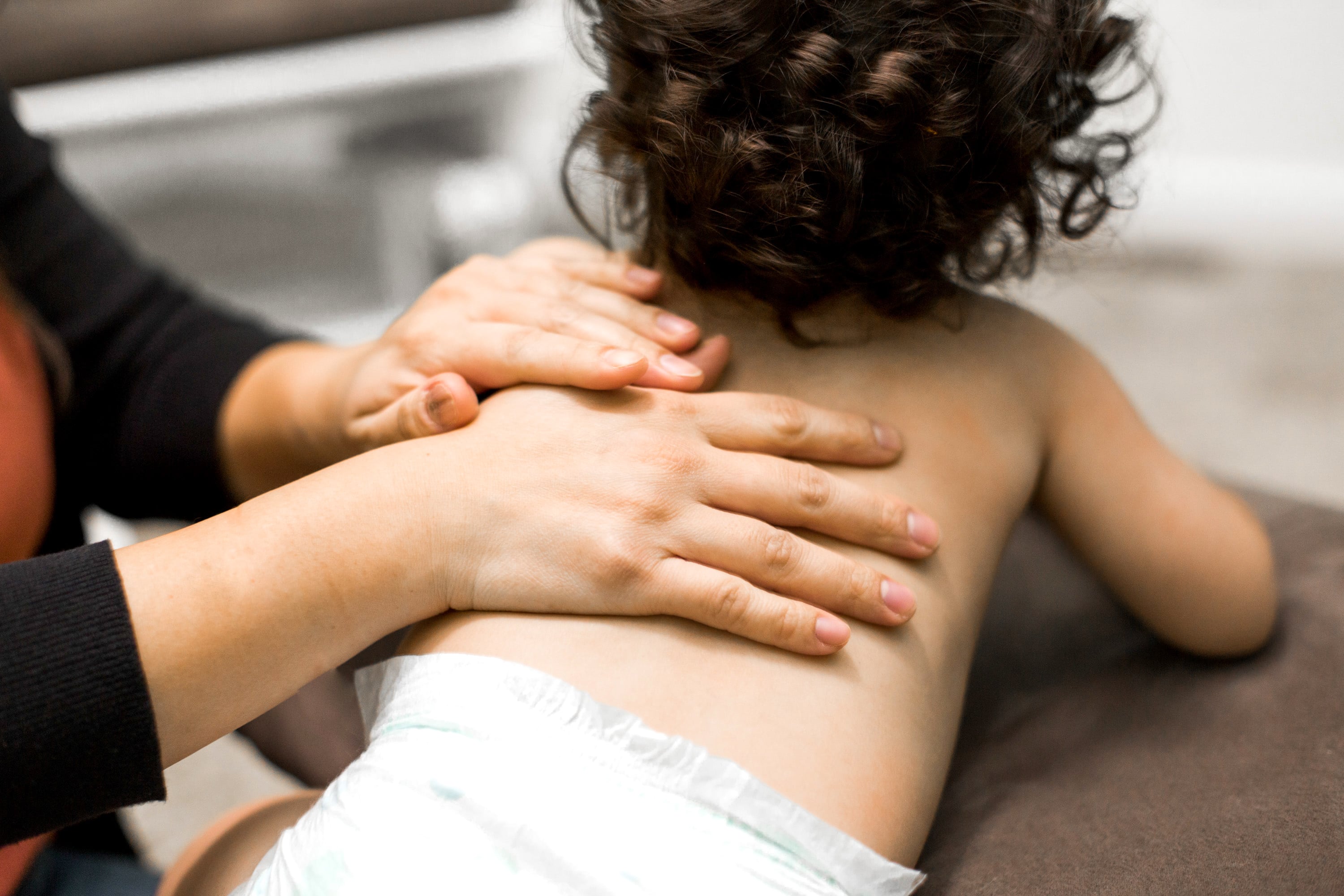 Un adulto está masajeando la espalda de un niño pequeño que usa pañal sobre una superficie acolchada en un entorno interior con luz suave.