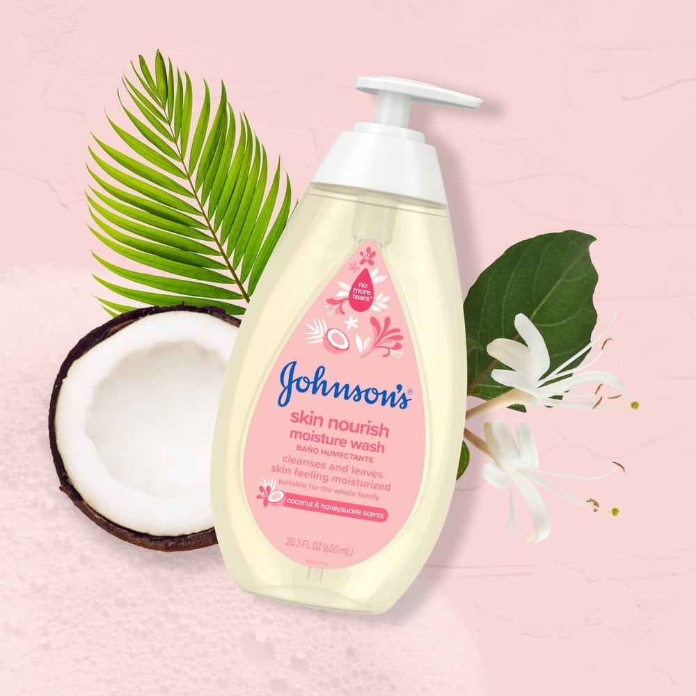 El envase de jabón líquido humectante Johnson's Skin Nourish descansa sobre un fondo rosa con un coco cortado por la mitad, hojas verdes y flores blancas. Texto: "limpia y deja la piel hidratada", y más.