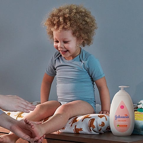 Un bebé feliz con cabello rizado sentado en un cambiador, vestido con un mameluco azul, con la mano de uno de los padres extendiéndose hacia él y un frasco de loción para bebé Johnson's cerca.