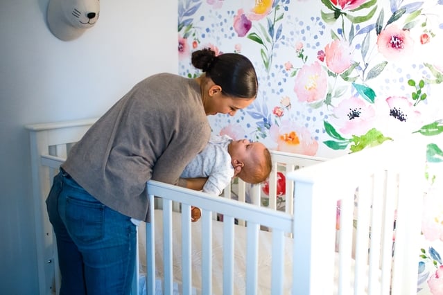 Una mujer se inclina sobre una cuna blanca, sonriendo a un bebé que está dentro. La habitación cuenta con un colorido mural floral y una cabeza de animal decorativa en la pared.