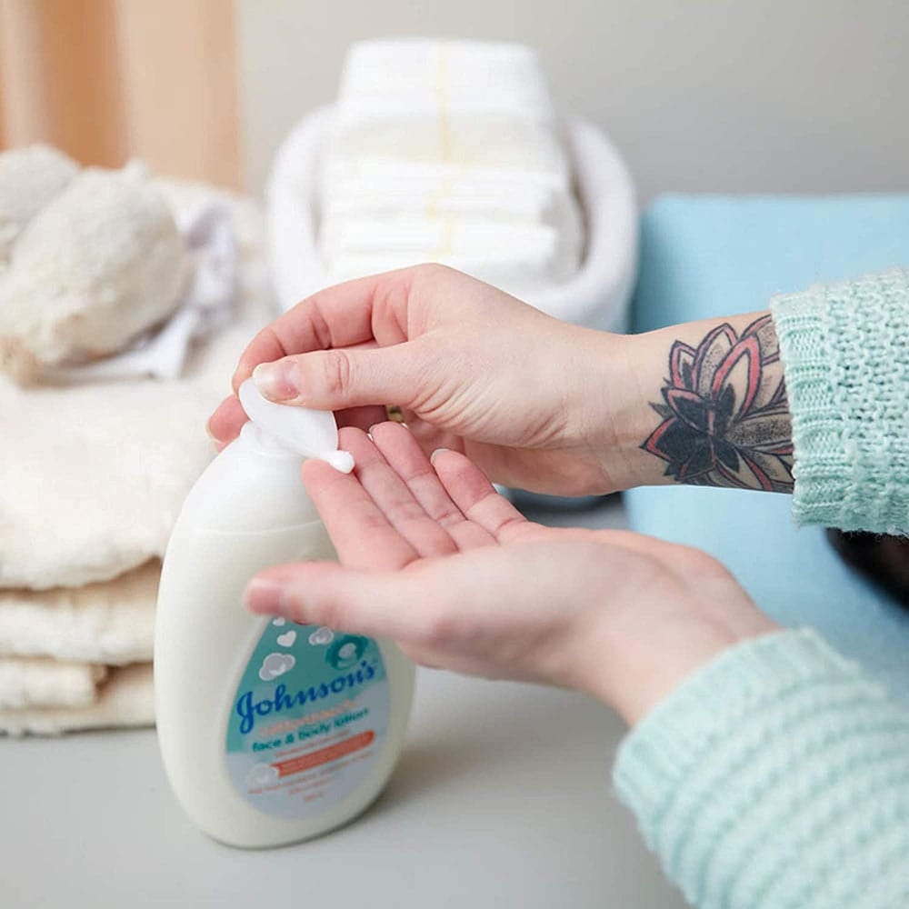 Persona dispensando la loción facial y corporal Johnson's Cotton Touch en sus manos, con artículos para el cuidado del bebé en el fondo.