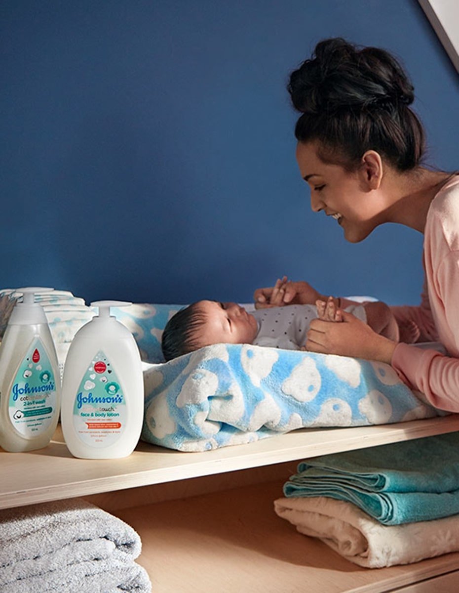 Una madre sonríe a su bebé, que está acostado en un cambiador con toalla azul estampada, junto a productos de baño para bebé Johnsons.