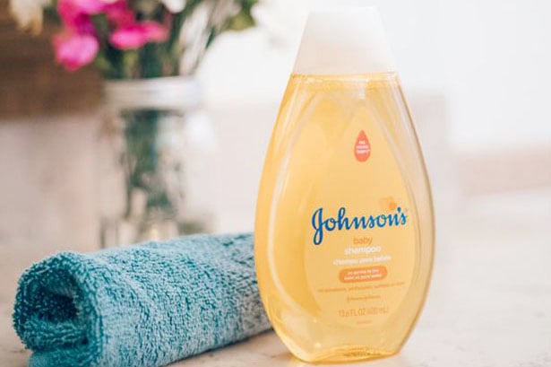 Champú Johnsons Baby, envase amarillo de 13,6 fl oz (400 ml) con tapa blanca, colocado sobre una encimera junto a una toalla azul, con flores en el fondo.