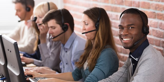 Representantes de atención al cliente en los escritorios, con auriculares y ayudando a las personas que llaman, uno de ellos sonriendo a la cámara.