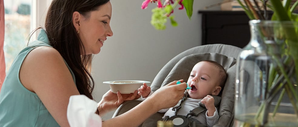 Una mujer alimenta a un bebé con una cuchara, sosteniendo un cuenco en una habitación moderna y bien iluminada con plantas y muebles al fondo.