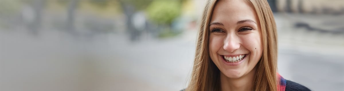 Una mujer sonríe francamente, su largo cabello cae alrededor de su rostro, en un entorno al aire libre con árboles y fondo borroso, lo que le da un ambiente alegre y vibrante.