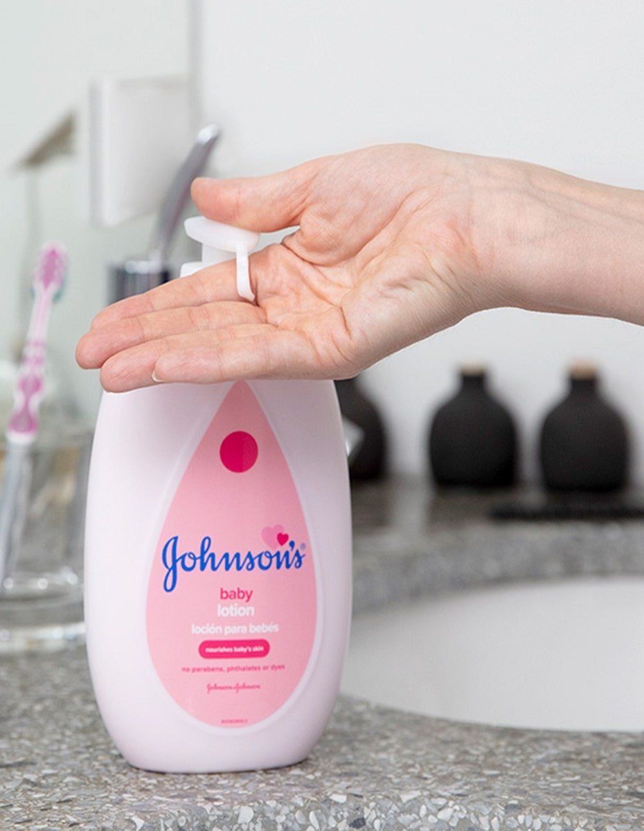 Una mano presiona la bomba de la loción Johnsons Baby, que está en un frasco rosa de 13,6 fl oz (400 ml) con un texto que indica "Clinically Proven Mildness" y "sin parabenos, ftalatos ni colorantes", en la encimera de un baño con cepillo de dientes y frascos en el fondo.