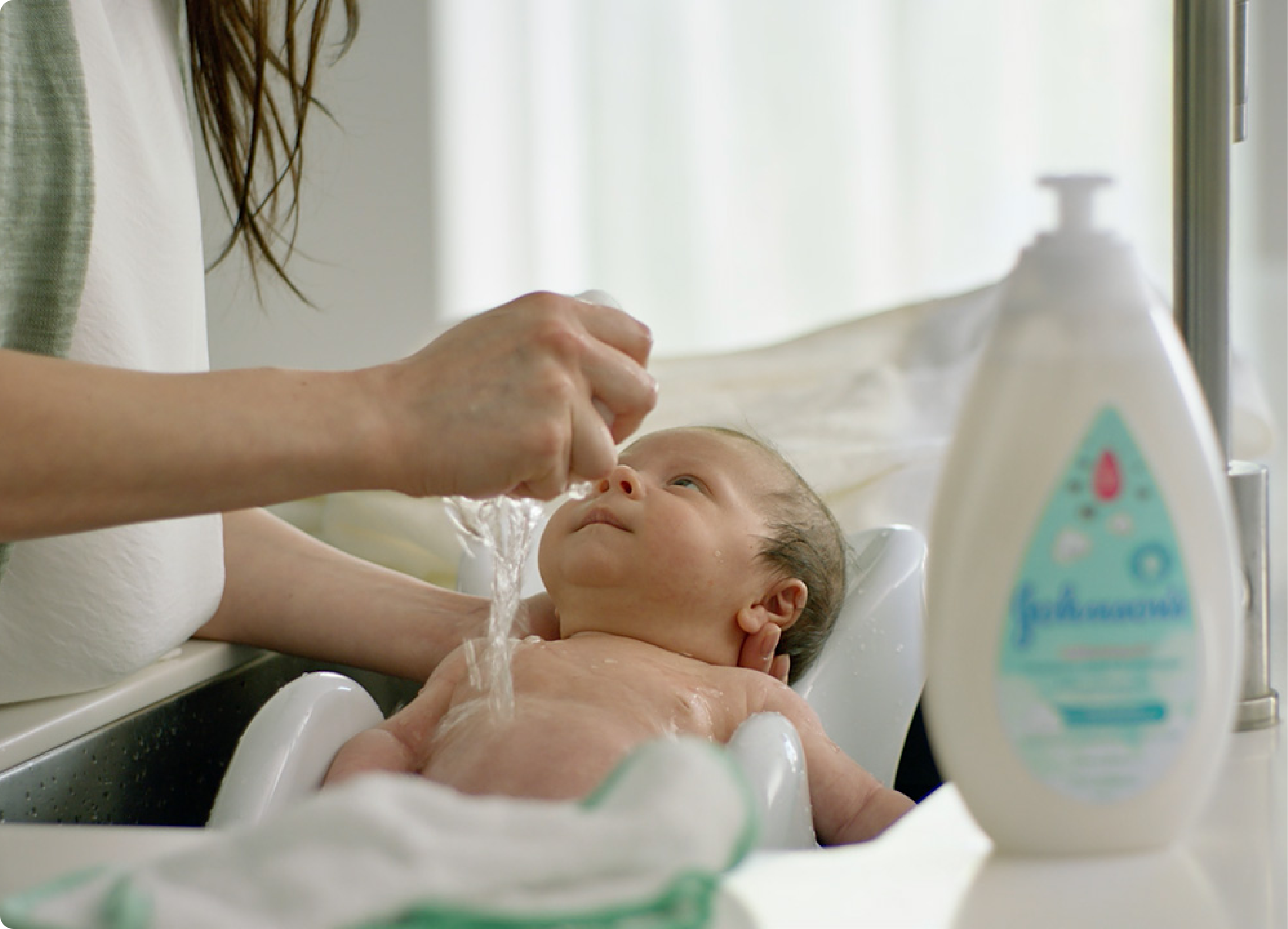 Un bebé es bañado en un lavabo por su madre, que vierte agua sobre su cabecita. Hay un envase de champú para bebé Johnson's Baby en el lado derecho de la imagen.