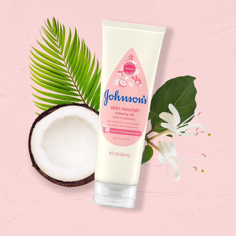 Un pomo de aceite cremoso de Johnson's Skin Nourish se encuentra cerca de un coco cortado por la mitad y hojas verdes sobre un fondo rosado. El texto en el pomo dice: "Aroma de coco y madreselva. 8 fl oz (236 ml)".