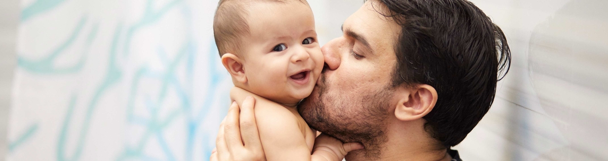 Un padre besando a su bebé que está en sus brazos, ambos con expresiones alegres.