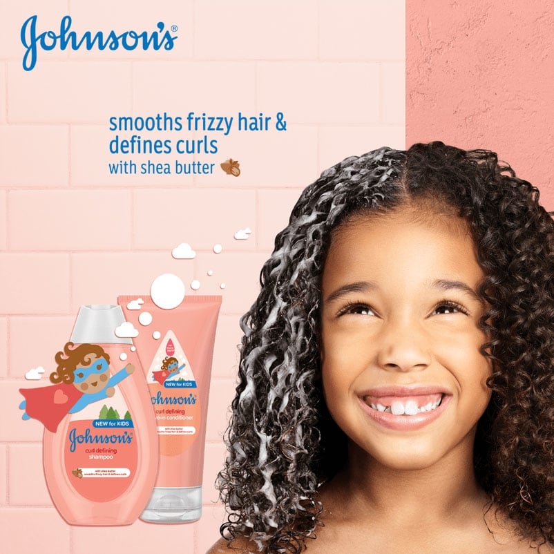 Chica sonriente con el pelo rizado cubierto de espuma, junto a los frascos de champú y acondicionador Johnson's Curl Defining con el texto "suaviza el cabello encrespado y define los rizos con manteca de karité".