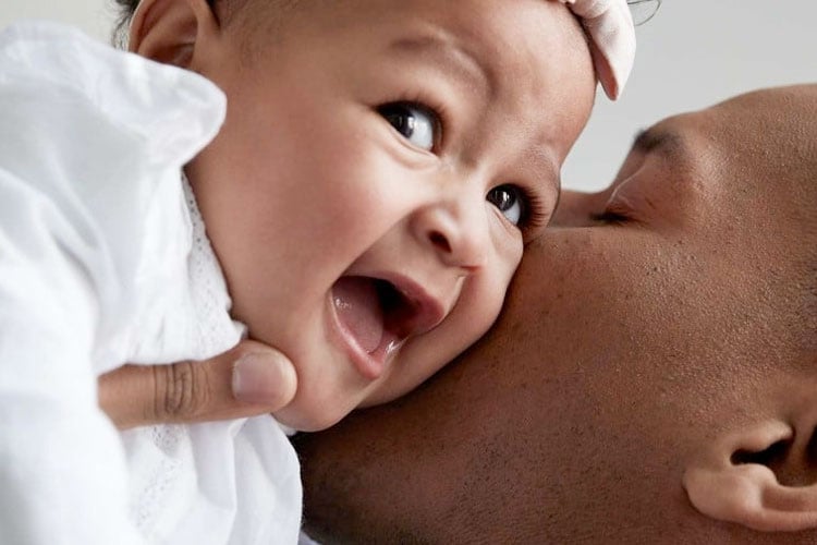 Un primer plano de un bebé sonriente siendo besado en la mejilla por un padre, que captura un momento alegre de afecto.