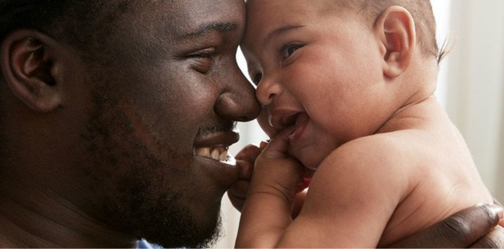 Un padre sonriente sosteniendo a su bebé feliz y tocando la nariz de este con la suya, ambos compartiendo un momento de unión y alegría.